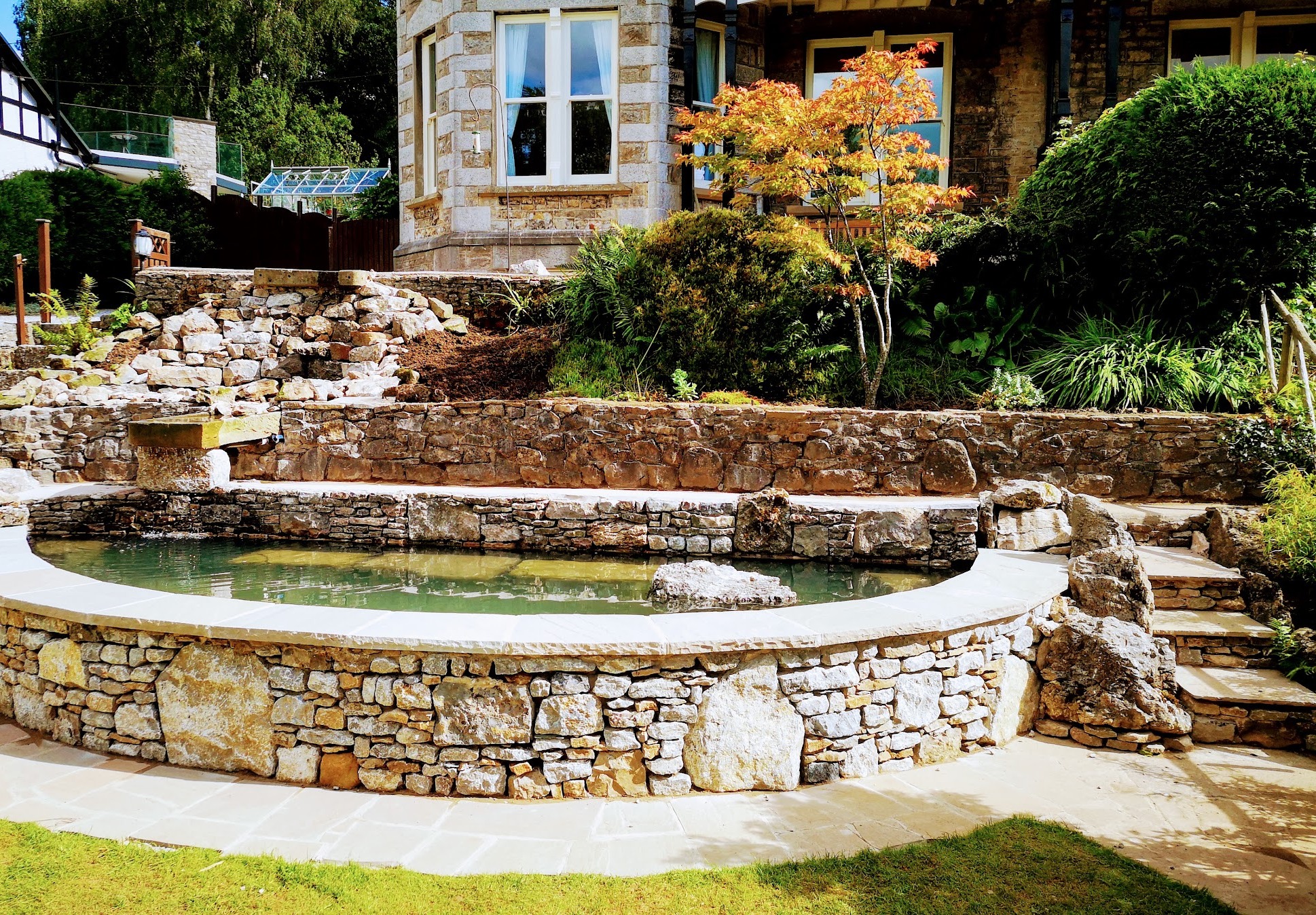Raised drystone-walled pond with stone steps and planting in front of a stone house