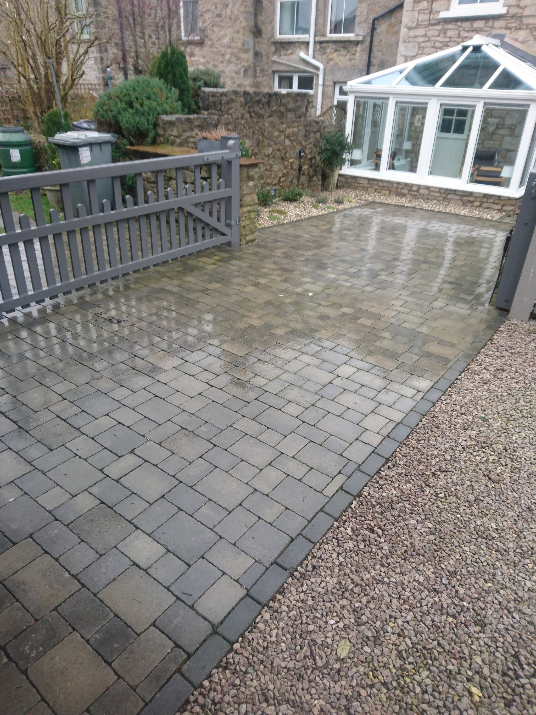 Block-paved yard with a grey gate in front of a stone house and conservatory